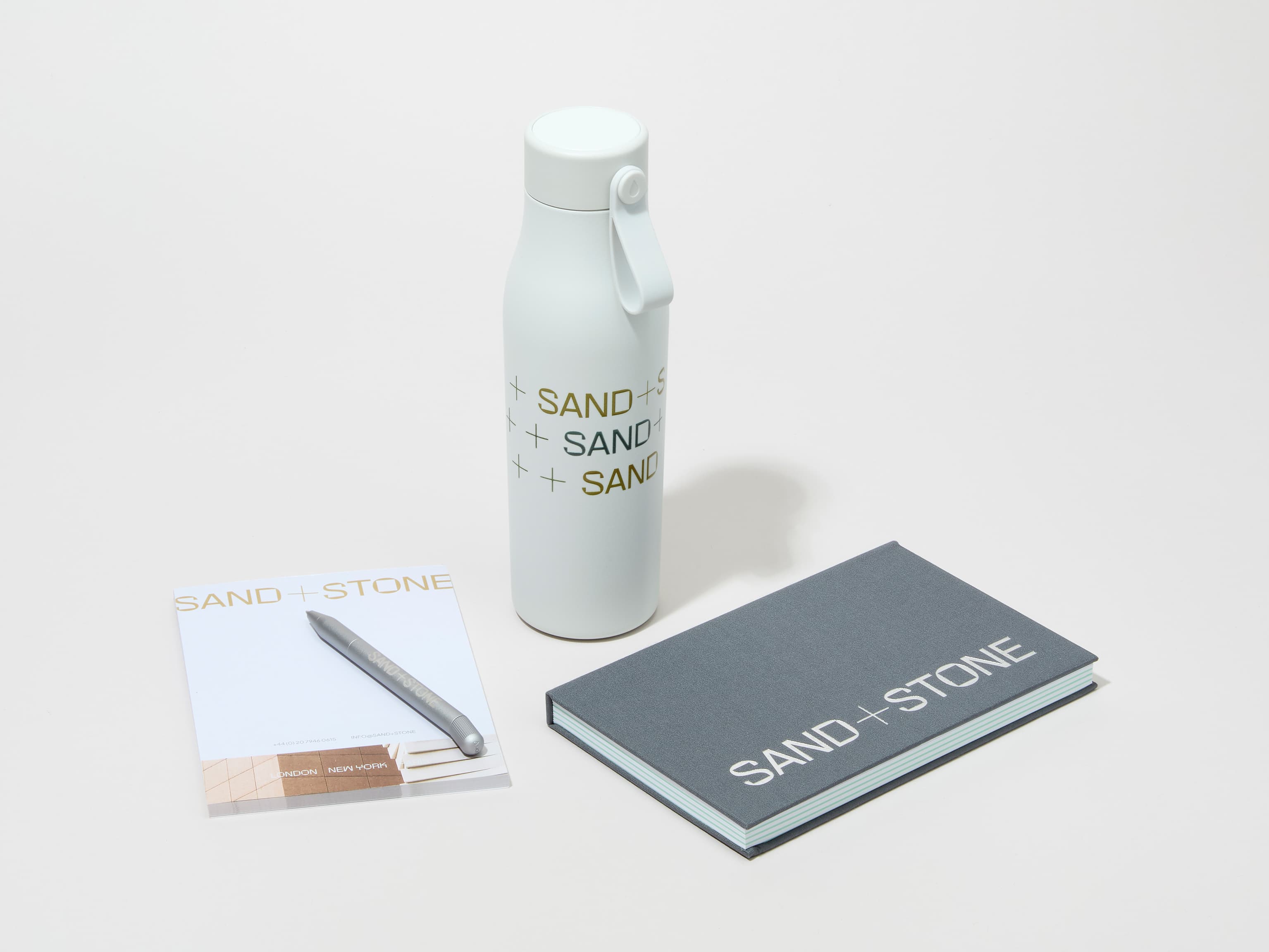 A collection of branded merchandise including a cloudy grey MOO water bottle with customized 360 wrap printing alongside a notepad, notebook and pen.