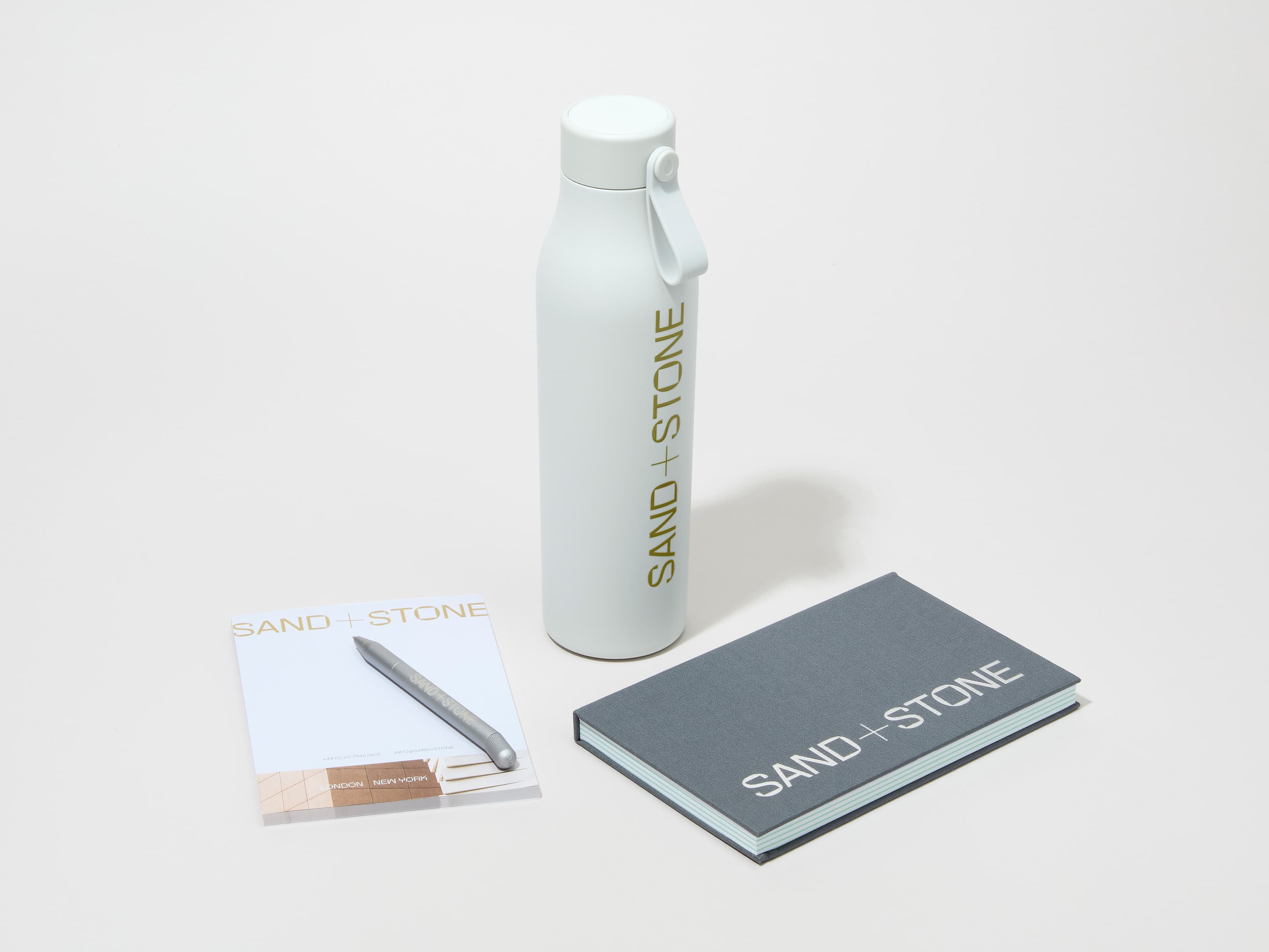 A collection image of a cloudy grey MOO water bottle with custom printing alongside a notebook, notepad and pen.