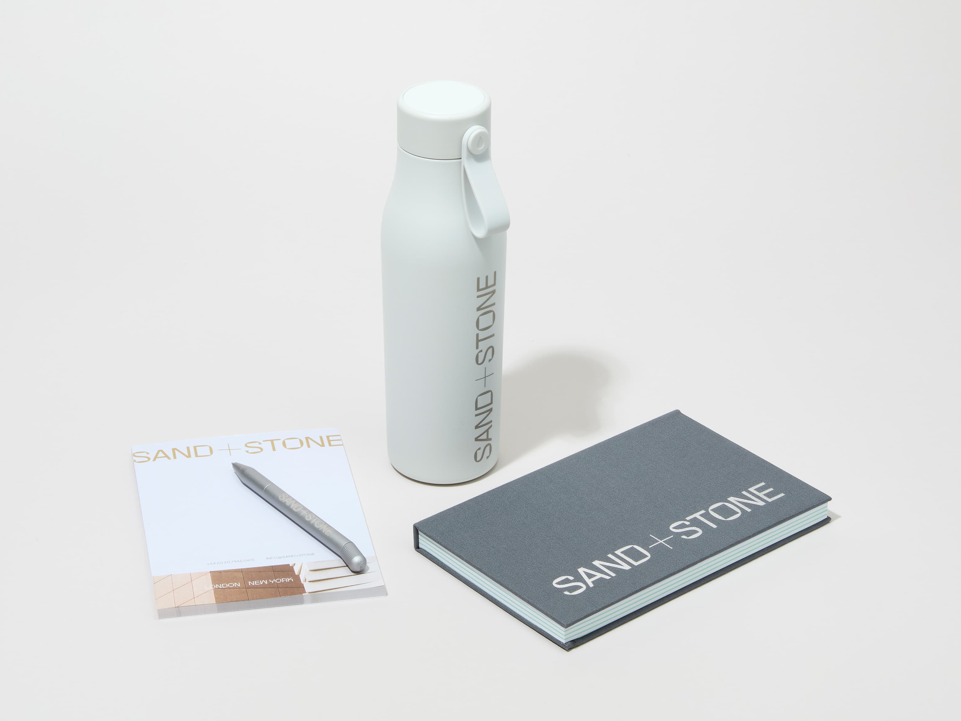 A collection of branded merchandise including a cloudy grey MOO water bottle with customized engraving alongside a notepad, notebook and pen.