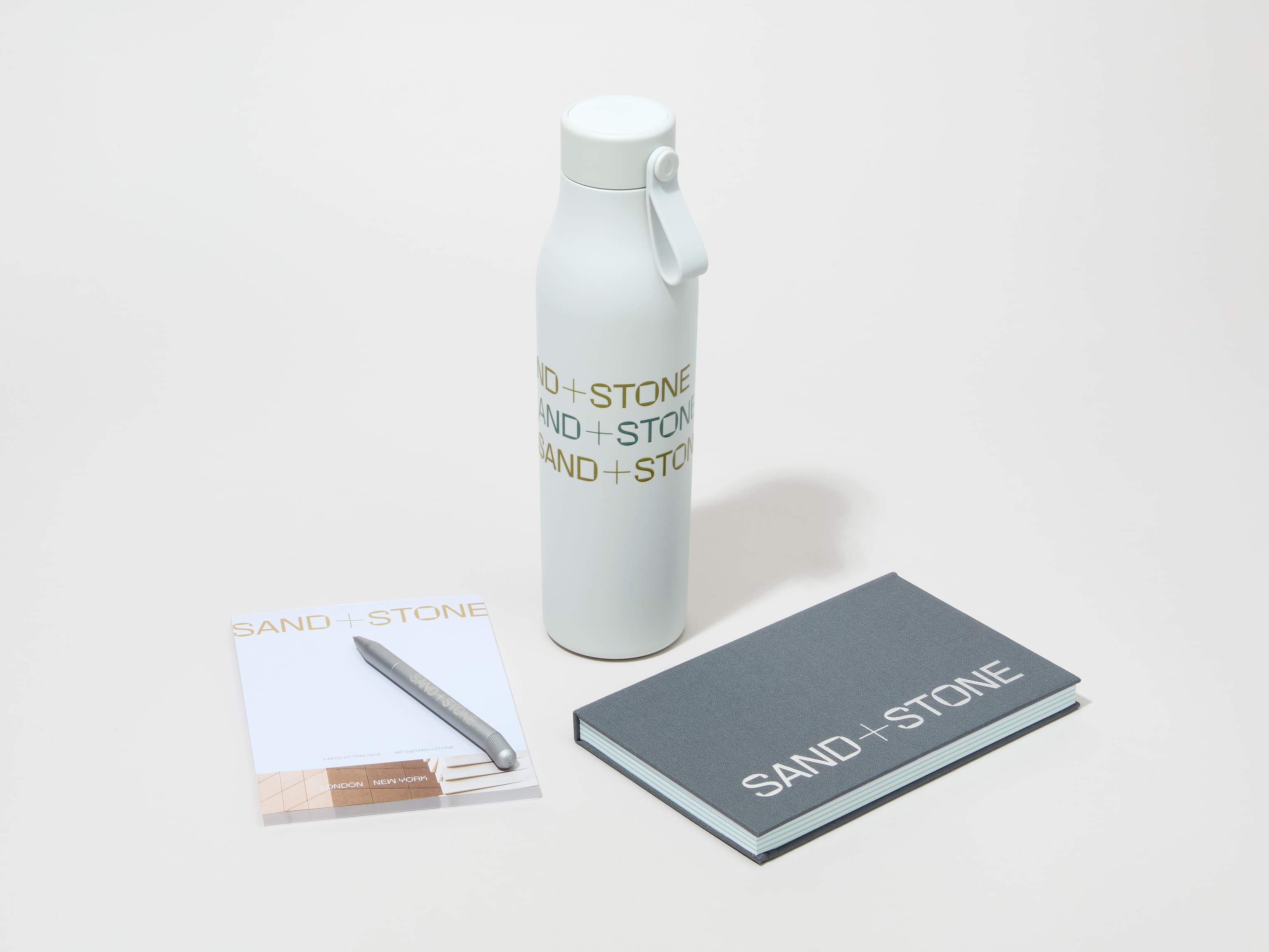 A collection image of a cloudy grey MOO water bottle with custom 360 wrap printing alongside a notebook, notepad and pen.