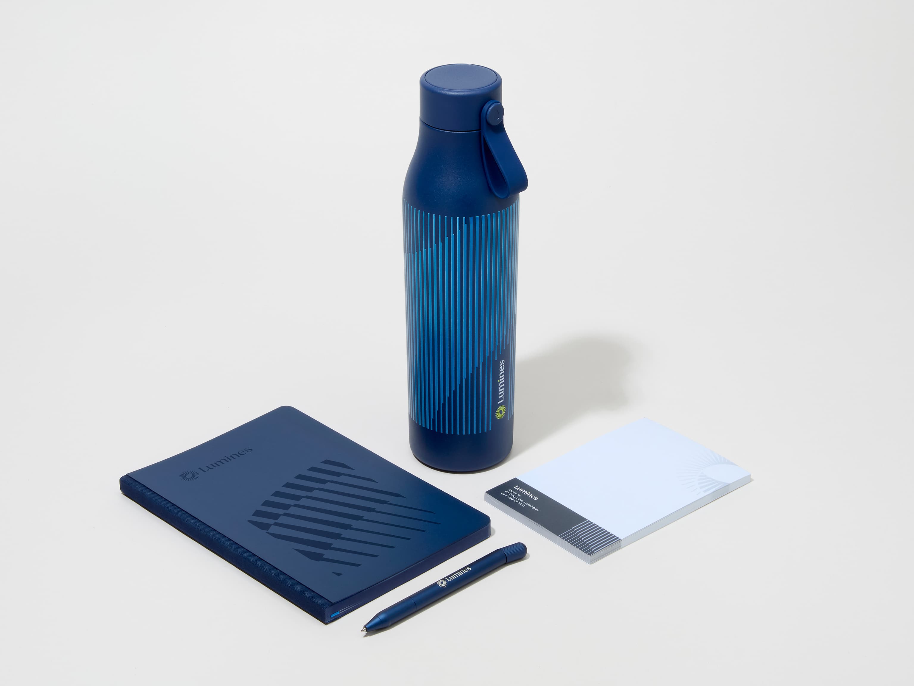 A collection image of a midnight blue MOO water bottle with custom 360 wrap printing alongside a notebook, notepad and pen.