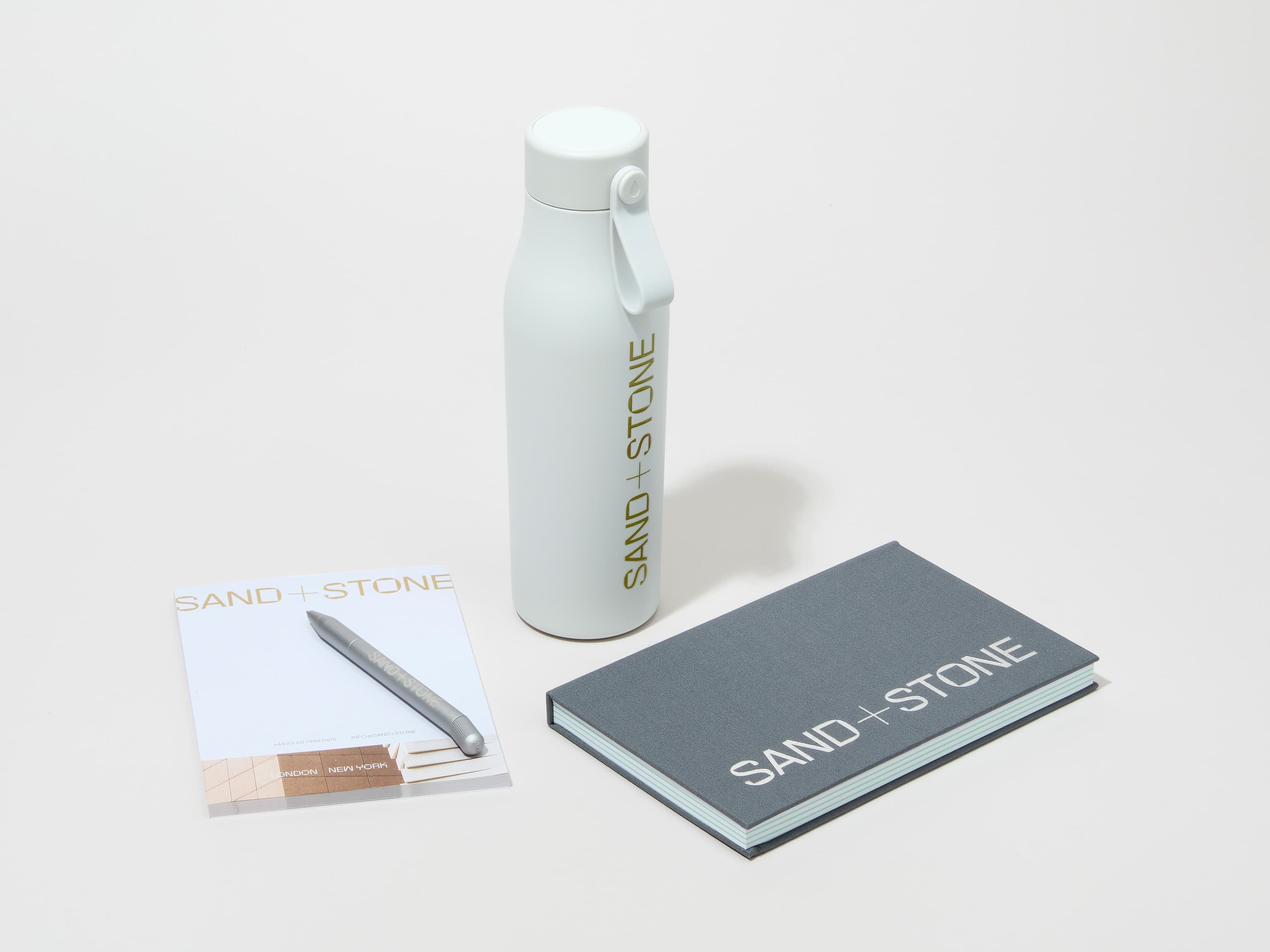 A collection of branded merchandise including a cloudy grey MOO water bottle with customized printing alongside a notepad, notebook and pen.