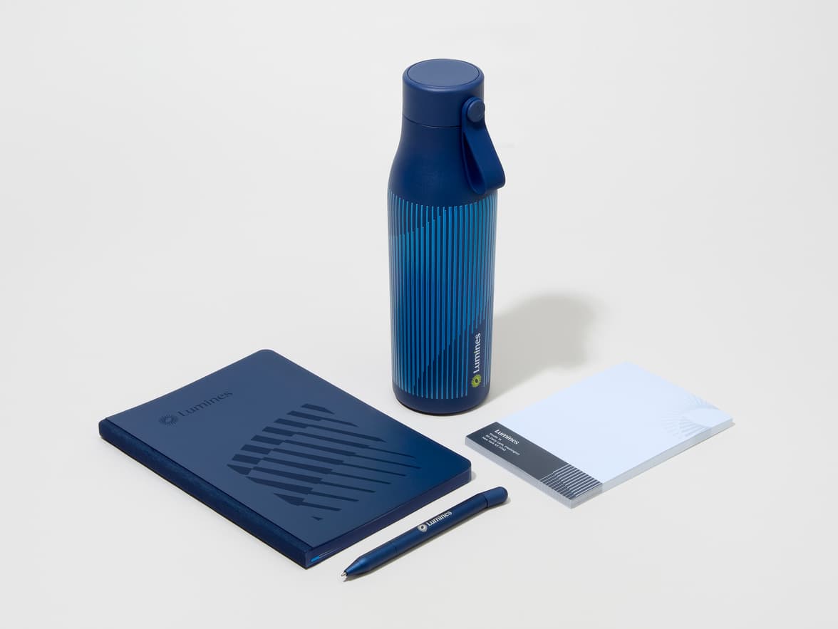 A collection of branded merchandise including a midnight blue MOO water bottle with customized 360 wrap printing alongside a notepad, notebook and pen.